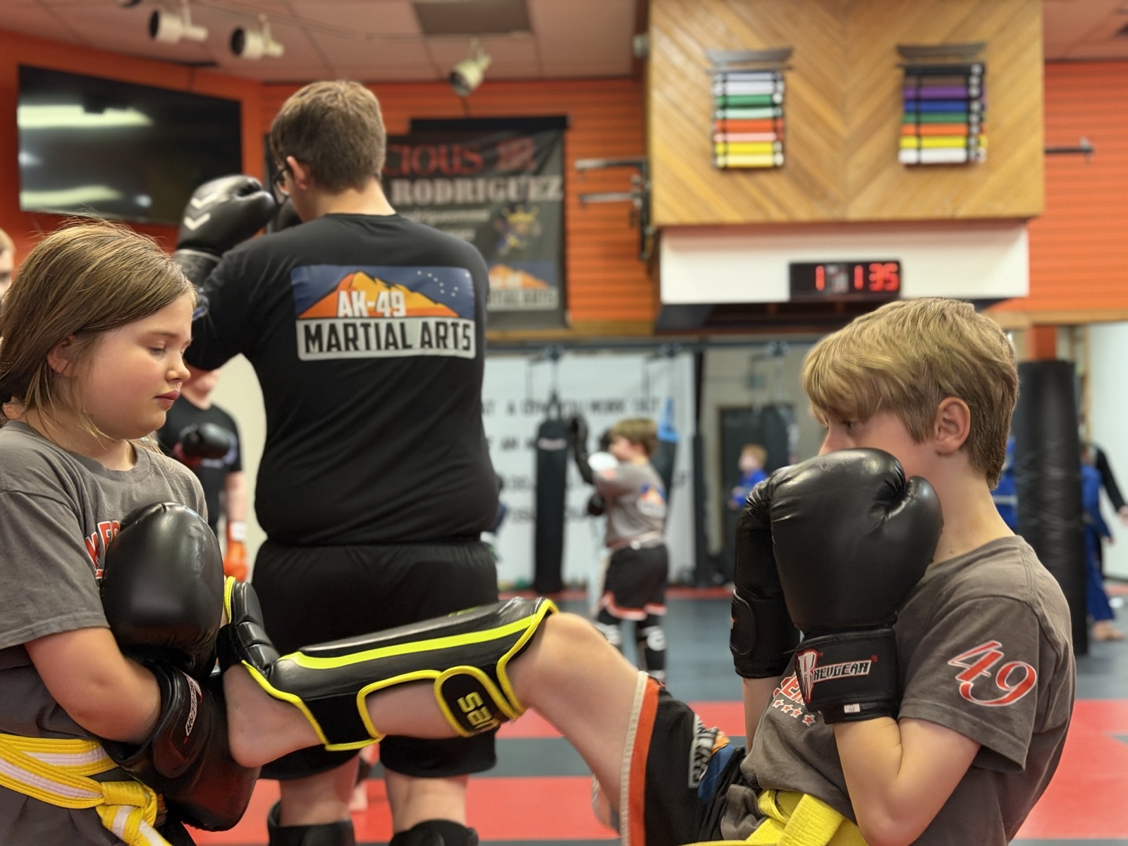 A kid in an AK-49 Martial Arts shirt kicking pads during a kids kickboxing class