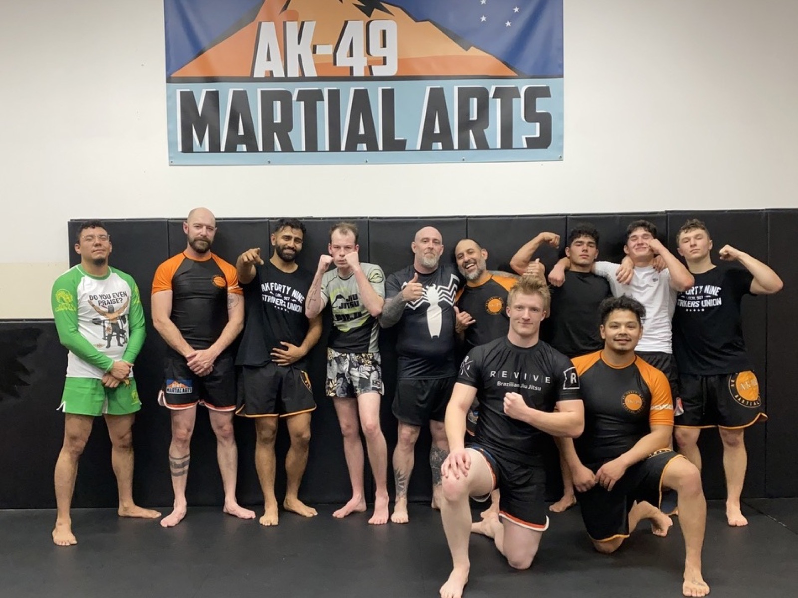 Adult students in fight shorts and rash guards standing together under the AK-49 Martial Arts banner