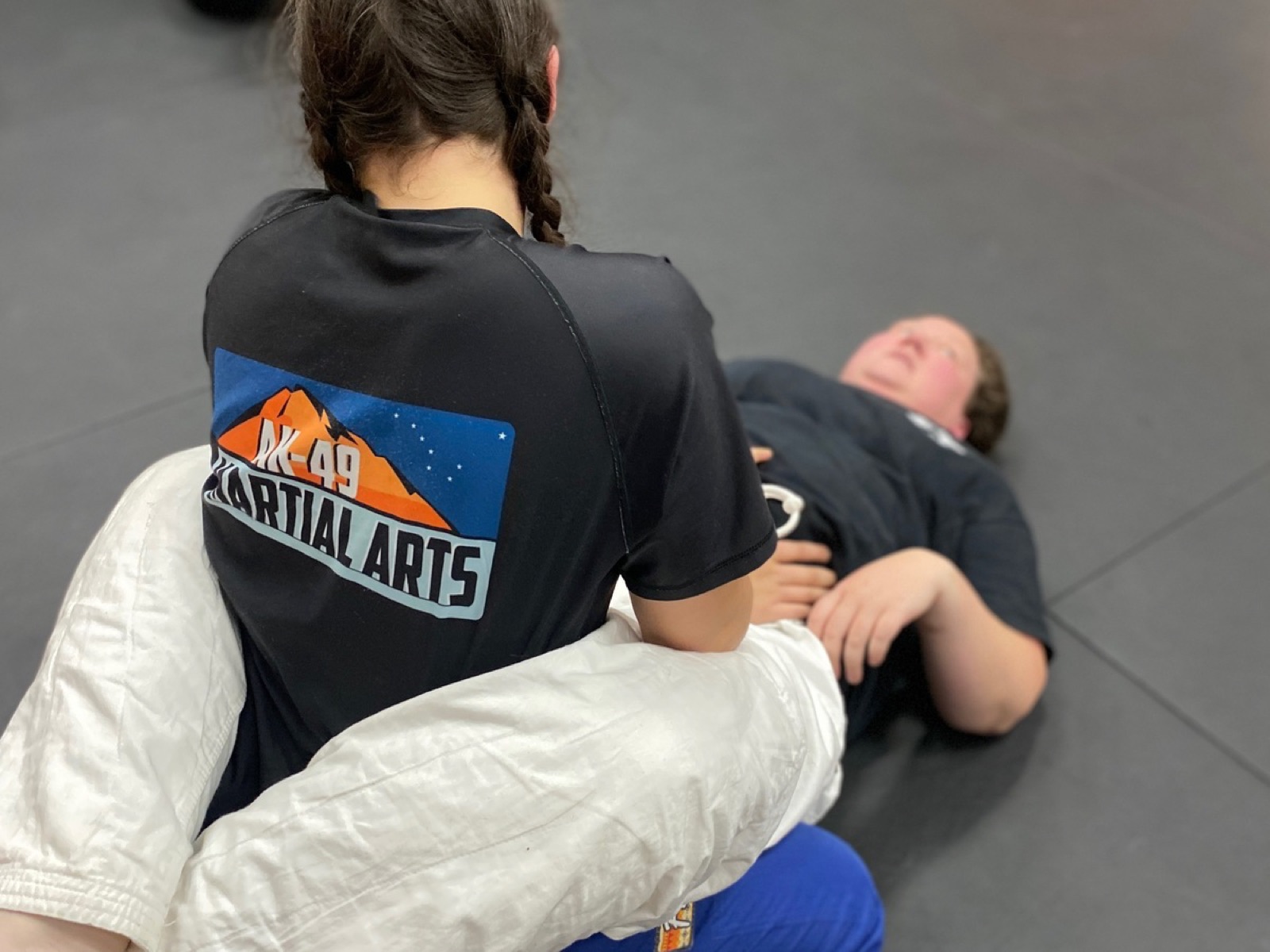 A student in an AK-49 Martial Arts t shirt and gi pants works a Jiu Jitsu position on the mat